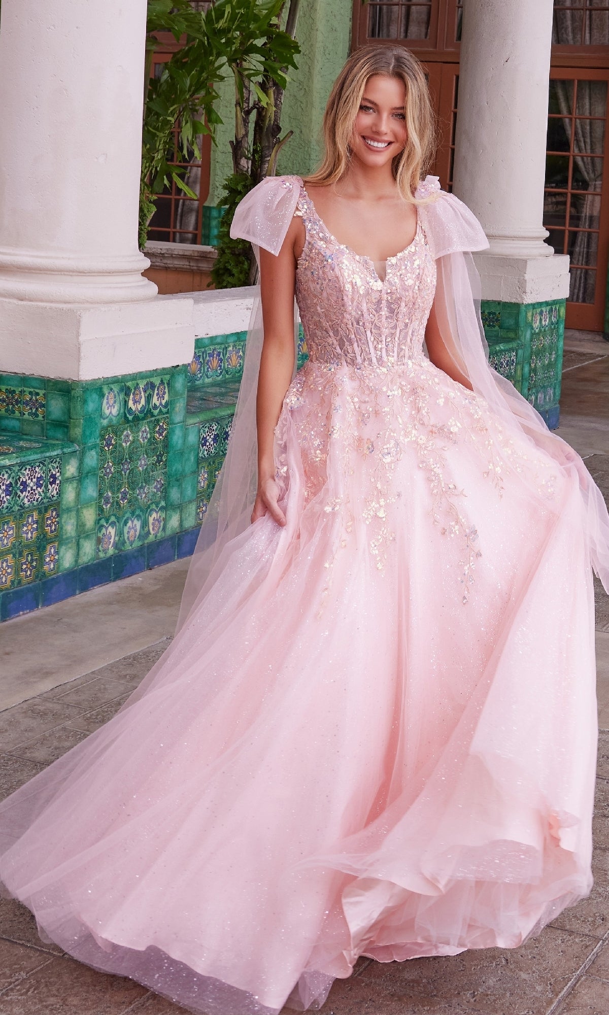 A woman in the Shoulder-Bows Long Prom Dress: Nox Anabel H1680—a sparkly light pink, floor-length gown with tulle sleeves—stands outdoors by tiled walls and white columns, smiling at the camera.